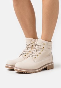 Bottines beiges en daim, avec des œillets dorés, des lacets épais et une semelle texturée. Un revêtement doux dépasse du haut.