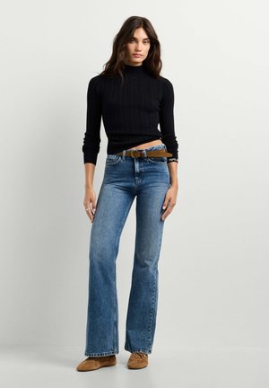 Donna che indossa un top nero a manica lunga, jeans blu a vita alta con cintura marrone e scarpe marroni, in piedi su uno sfondo chiaro uniforme.