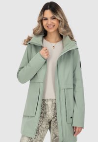Hellgrüne, wasserdichte Jacke mit Reißverschluss, Fronttaschen und Kapuze. Tragen über einem cremfarbenen Oberteil und texturierten Schlangenhautmuster-Hosen.