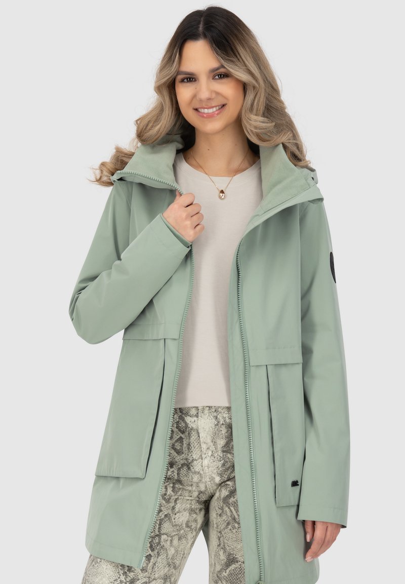 Hellgrüne, wasserdichte Jacke mit Reißverschluss, Fronttaschen und Kapuze. Tragen über einem cremfarbenen Oberteil und texturierten Schlangenhautmuster-Hosen.