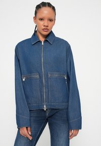Giacca di jeans di colore blu scuro, con zip frontale, due tasche sul petto con cerniera, maniche larghe e cuciture a contrasto.