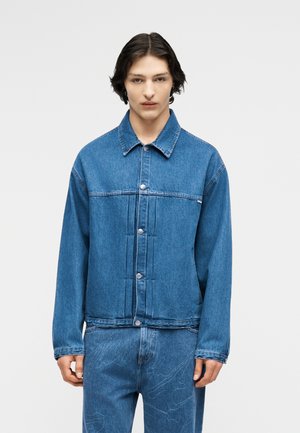 Noor keskmise pikkusega tumedate juustega, seljas nööbitud sinist teksajopet ja sobivad sinised teksad peente mustritega.