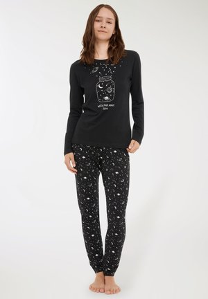Donna in pigiama nero con motivi celestiali bianchi, una grafica di un barattolo sulla parte superiore e la scritta "Guarda la tua magia crescere," in piedi a piedi nudi.