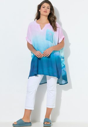 Kobieta w luźnej pastelowo różowo-niebieskiej tunice ombre z przednią kieszenią, białych krótkich spodniach i niebieskich klapkach, stojąca na tle białej ściany.