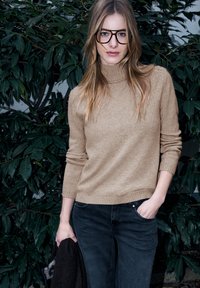 Beiger Rollkragenpullover mit geripptem Saum und Bündchen, kombiniert mit dunkler Jeans. Das Modell hält eine dunkle Jacke vor einem Hintergrund aus grünen Blättern.