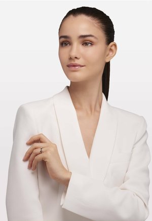 Blazer bianco con un profondo scollo a V. La modella mostra un anello sulla mano sinistra, con una gemma centrale e accenti sottili.