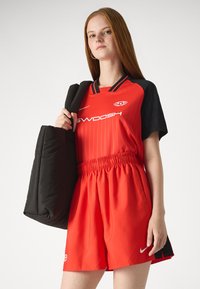 Červený športový dres s čiernymi rukávmi a prúžkami, kombinovaný s červenými šortkami. Model drží čiernu tašku s textúrovaným povrchom.