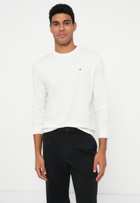 Homem vestir uma camisa de mangas compridas branca e calças pretas, de pé contra um fundo branco liso, olhando em frente com uma expressão neutra.