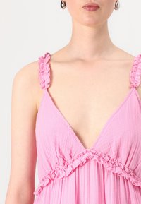 Sleeveless pink dress with ruffled straps and a deep V-neckline. Soft, textured fabric with gathered detailing at the waist.