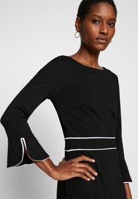 Abito nero con maniche a campana lunghe, con bordature bianche sulle maniche e in vita. La texture liscia e il design aderente mettono in risalto la silhouette.