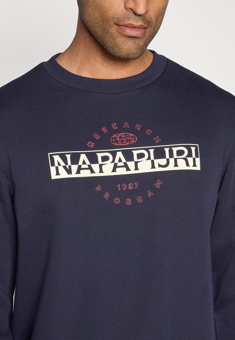 Homme portant un sweat-shirt bleu marine avec le logo "NAPAPIJRI" et un texte circulaire rouge "PROGRAMME DE RECHERCHE 1987" sur la poitrine.