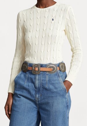 Persoon in een romige kabelgebreide trui met blauwe geborduurde logo, hoge blauwe spijkerbroek en een beige riem met grote zilveren conchos.