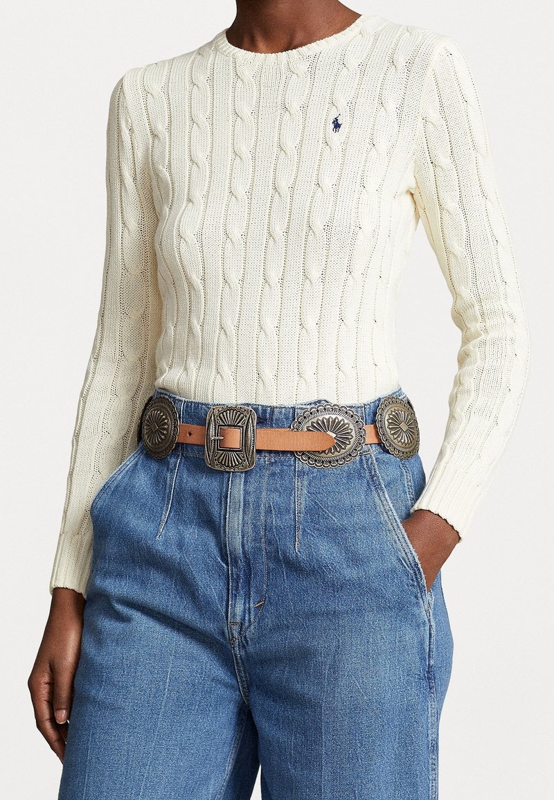 Krämfärgad kabelstickad tröja med rund halsringning, kombinerad med högmidjade jeans och ett brunt läderbälte med silverfärgade dekorativa spännen.