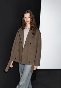 Double-breasted, olive green jacket with black buttons, oversized fit, and side tie detail, worn over a gray top and light blue jeans.