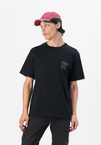 Camiseta negra de manga corta con cuello redondo y pequeño logotipo impreso en el lado izquierdo del pecho. El modelo lleva una gorra rosa, combinada con jeans oscuros.