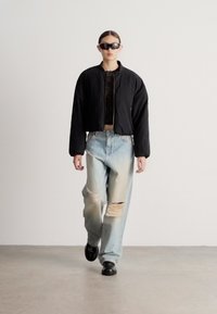 Čierna bomber bunda cez pruhované tričko, doplnené svetlomodrými roztrhanými džínsami s rozparkami. Doplnky zahŕňajú tmavé slnečné okuliare a čierne topánky.