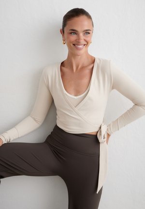 Femme souriante portant un haut beige cache-cœur et des leggings foncés, posant avec une jambe pliée sur un fond blanc uni.