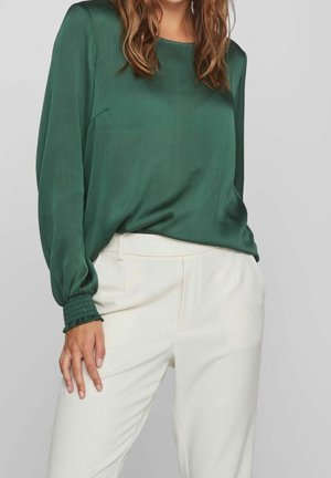 Femme portant un chemisier vert foncé à manches longues avec des poignets smockés et un pantalon taille haute écru, debout devant un fond uni.