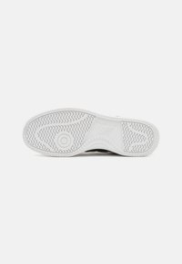 Talpa de sneaker neagră cu talpă exterioară din cauciuc alb, având un model de profil ondulat și un accent circular. Logo-ul "New Balance" embosat pe lateral.