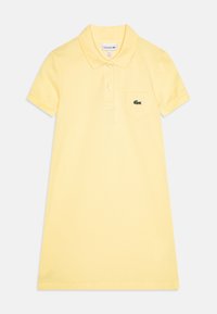 Polodress i ljusgul bomull med korta ärmar, krage och ett bröstfick med en grön Lacoste-logga.