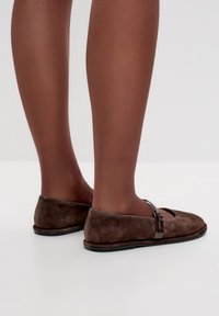 Ballerine in suede marrone con punta rotonda, cinturino sulla parte superiore del piede e suola nera. Sono visibili gambe che indossano collant trasparenti marroni.