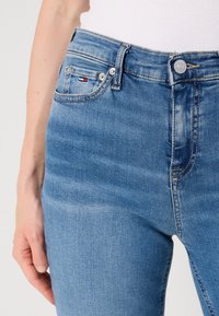 Jean en denim bleu clair avec une coupe slim, présentant un design classique à cinq poches, une fermeture à bouton en métal et des détails d'usure subtils.