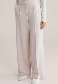 Žena u svijetloružičastim širokim hlačama, bijeloj teksturiranoj majici s dugim rukavima i bijelim tenisicama, stoji s jednom rukom u džepu.