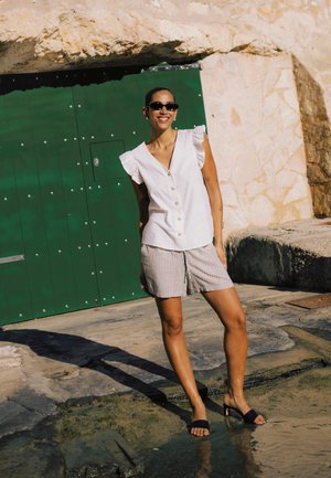 Ženska stoji na prostem v črtastih kratkih hlačah, belem brezrokavniku z gumbi, črnih sandalih z visokimi petami in sončnih očalih ob kamniti steni z zelenimi vrati.
