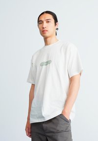 Camiseta de color gris claro con escote redondo y mangas cortas, que presenta un diseño gráfico verde en el centro. Tejido de algodón suave.