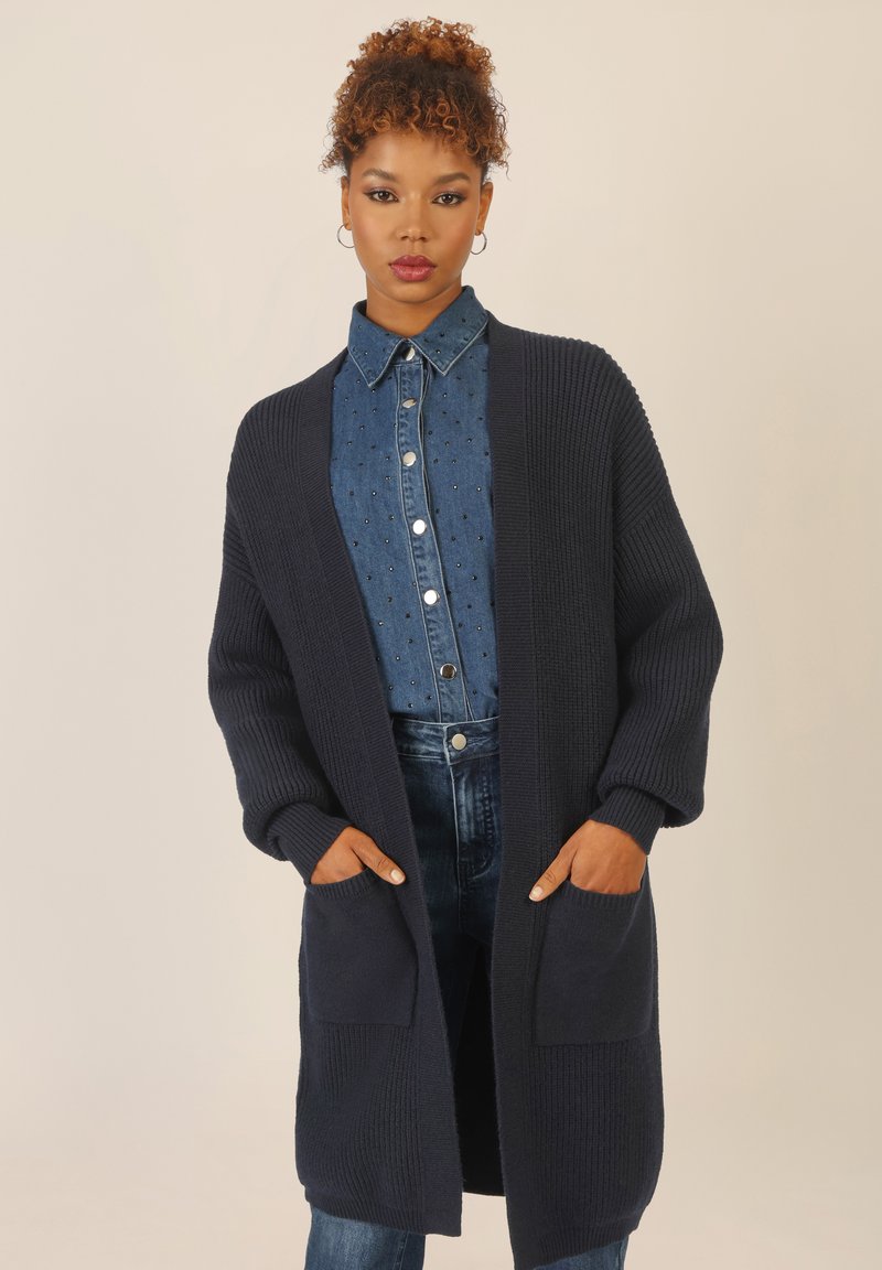 Granatowy długi kardigan wełniany o ribbowanej fakturze, z otwartym przodem i dwiema przednimi kieszeniami, noszony na koszuli z denimy z guzikami oraz dżinsach.