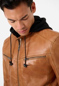 Braune Lederjacke mit schwarzer Kapuze, ausgestattet mit Reißverschlusstaschen auf der Brust und Zugbändern. Glatte Oberfläche mit minimalen Nähdetails.