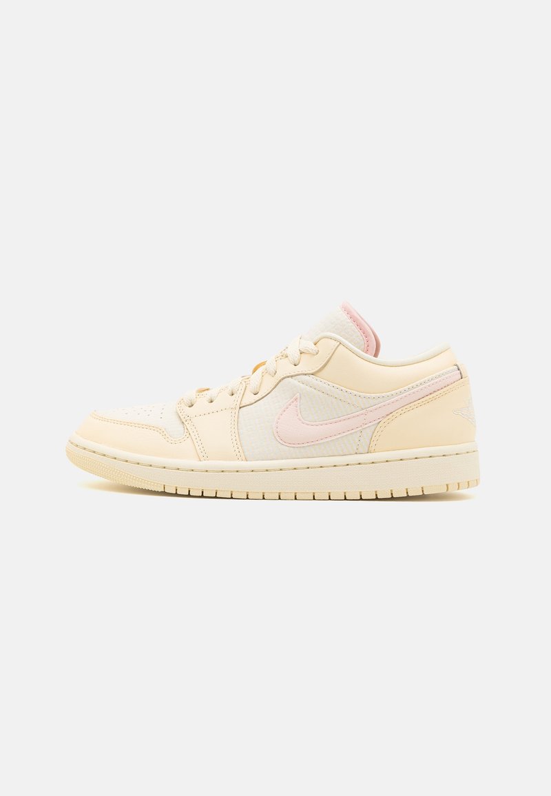 Baskets basses crème avec swoosh Nike rose pâle et détails, boîte à orteils perforée, semelle en caoutchouc sur un fond clair uni.