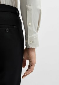 Witte overhemdd met kleine, gestructureerde stippen, met knopen bij de manchetten; gecombineerd met zwarte, gestructureerde broek. Een hand is aan de zijkant iets zichtbaar.