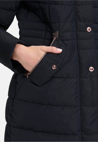 Cappotto imbottito blu navy con cuciture a quadri orizzontali, dotato di una tasca laterale con zip e hardware in oro rosa e un accento triangolare in tessuto marrone.