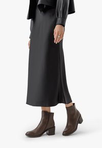 Kvinna i lång svart kjol, svart läderjacka och bruna läderankelboots med klackar, står framför en vit bakgrund.