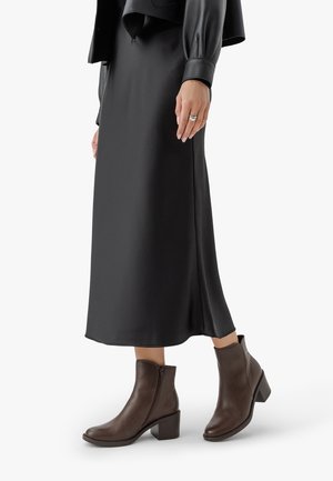 Vrouw gekleed in een lange zwarte rok, een zwarte leren jas en bruine leren enkellaarsjes met blokhakken, staande tegen een witte achtergrond.
