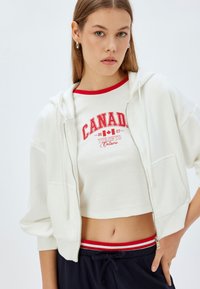 Hvid zip-hoodie med en afslappet pasform og korte ærmer, båret over en cropped hvid skjorte med rød Canada grafisk tekst og detaljer.