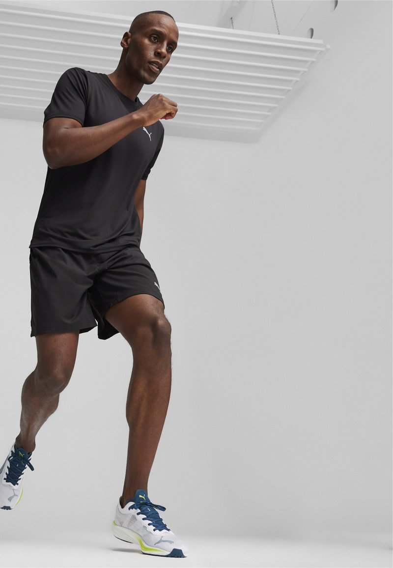 T-shirt sportivo nero e pantaloni corti, abbinati a scarpe da corsa bianche e blu con dettagli verdi, modello in posizione di corsa.