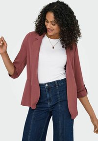 Kompakte, leichte burgunderfarbene Blazer mit offenem Vorderteil, dreiviertellangen Ärmeln und glatter Textur, kombiniert mit einem weißen Hemd und Jeans.
