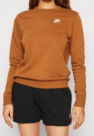 Femme portant un sweat-shirt Nike marron et un short Nike noir avec des logos sur la poitrine et la jambe gauche, debout, les bras détendus le long du corps.