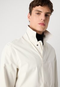 Chaqueta blanca impermeable con un alto cuello, cremallera frontal y botones a presión. Presenta una textura suave y elegante con un diseño minimalista.