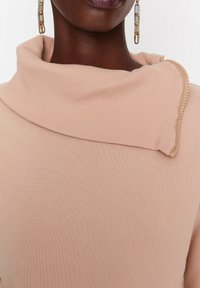 Top de punto acanalado rosa claro con cuello de cisne y un detalle de cremallera dorada en el cuello. La tela parece suave con una textura lisa.