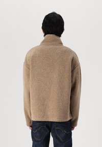 Beige, fluffy fleece jack met een hoge kraag, relaxte pasvorm en lange mouwen, gecombineerd met donkere spijkerbroeken, bekeken vanaf de achterkant.