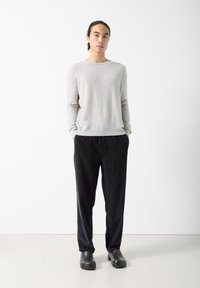 Mladić s dugom tamnom kosom vezanom unazad, obučen u svijetlosivi džemper, crne manšestarske hlače i crne papuče bez vezica, stoji na bijeloj pozadini.