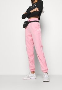 Světle růžové tepláky s elastickými manžetami, bílými bočními pruhy a vyšitým logem. Spárované s černým topem s dlouhým rukávem a bílými teniskami.