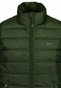 Grüne Puffjacke mit hohem Kragen, frontalem Reißverschluss und gestepptem Innenfutter. Trägt den Markennamen „GANT“ auf der linken Brust. Strukturierte Oberfläche.