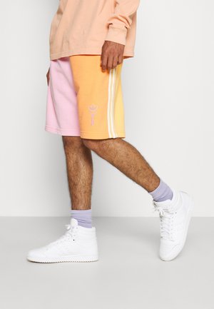 Person iført pastelrosa og orange shorts med hvide striber, lyse lilla sokker og hvide high-top sneakers, stående mod en ensfarvet baggrund.