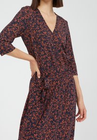 Robe florale marine à manches trois-quarts, décolleté en V, taille cintrée et légère finition à nouer sur le côté. Présente de petites fleurs orange.
