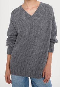 Pull gris côtelé avec col en V, coupe décontractée et manches retroussées, fabriqué en maille douce et texturée. Associé à un jean bleu clair.