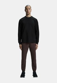 Camicia nera a maniche lunghe con tasca frontale, abbinata a pantaloni tapered marrone scuro e scarpe da ginnastica nere. Design minimalista e vestibilità rilassata.